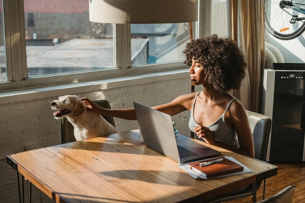 Young woman working at home on laptop, petting her dog. Sunlit room with modern decor and a warm vibe.