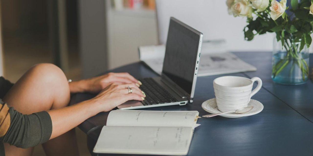Female freelancer using laptop with coffee at home office desk, surrounded by roses and a planner.